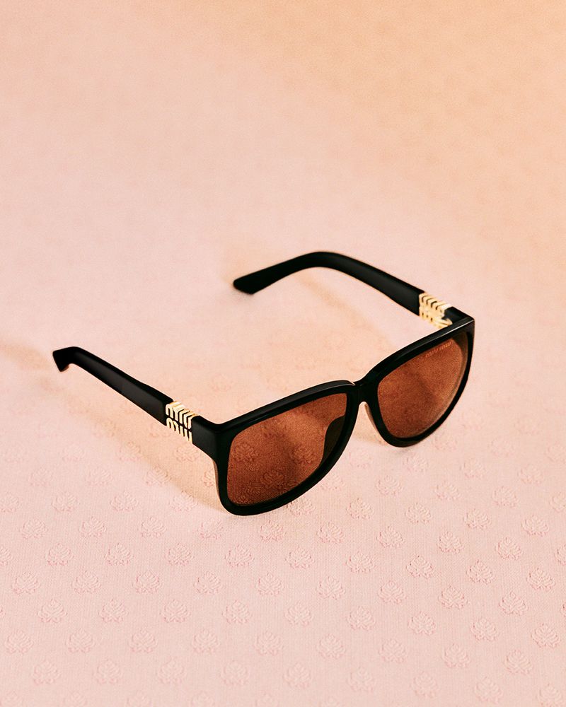 Studio shot of black framed sunglasses with brown lenses, accented with metallic gold, resting on a light pink patterned background.