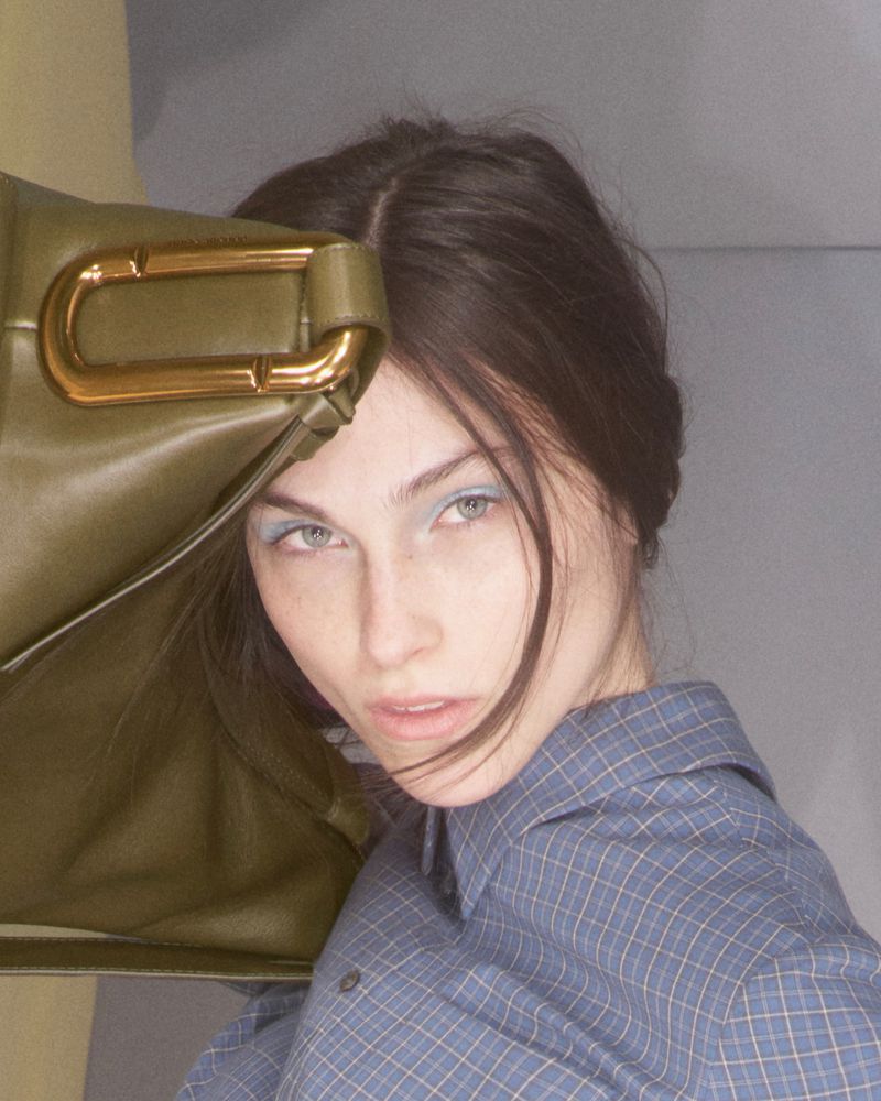 Close-up portrait of a woman in a blue plaid collared shirt posing beside an olive green leather bag with gold hardware.