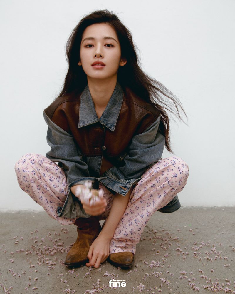 A young Asian woman in casual clothing, crouches indoors with flowers in her hands, against a plain background, in a fashion editorial style.