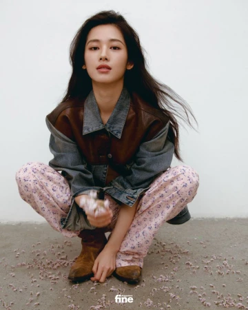 A young Asian woman in casual clothing, crouches indoors with flowers in her hands, against a plain background, in a fashion editorial style.
