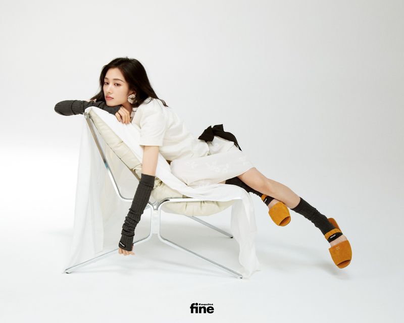 A stylish woman reclines in a modern chair, sporting a white dress, arm warmers, socks, and slip-on shoes in a clean studio setting.