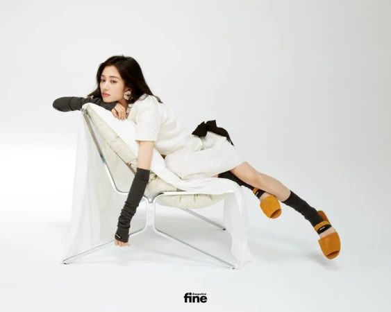 A stylish woman reclines in a modern chair, sporting a white dress, arm warmers, socks, and slip-on shoes in a clean studio setting.