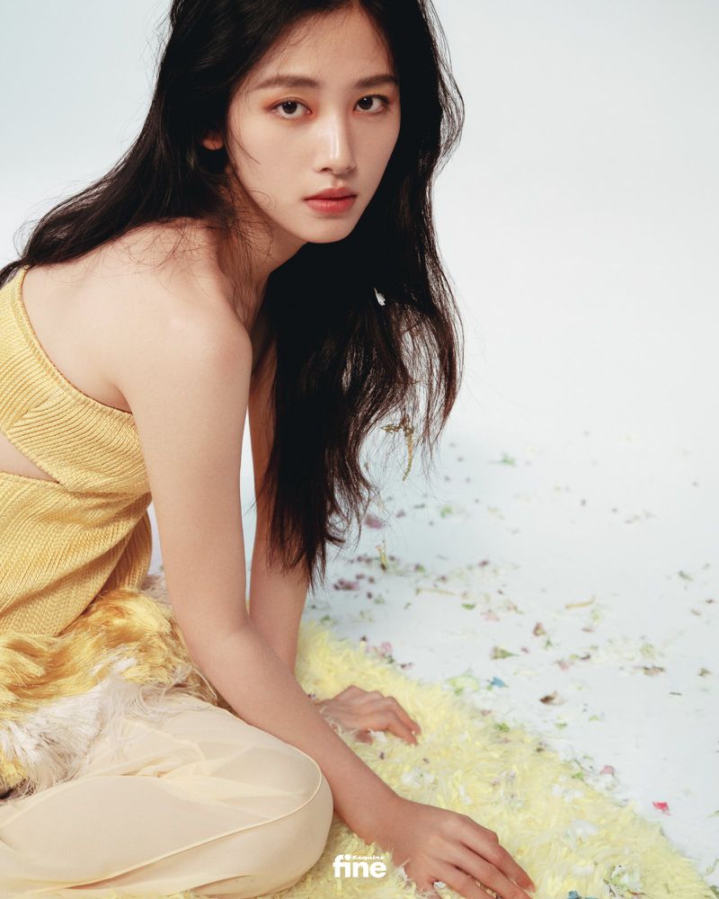Portrait of a young Asian woman with long dark hair, wearing a gold top and pale pants, posing against a neutral background.