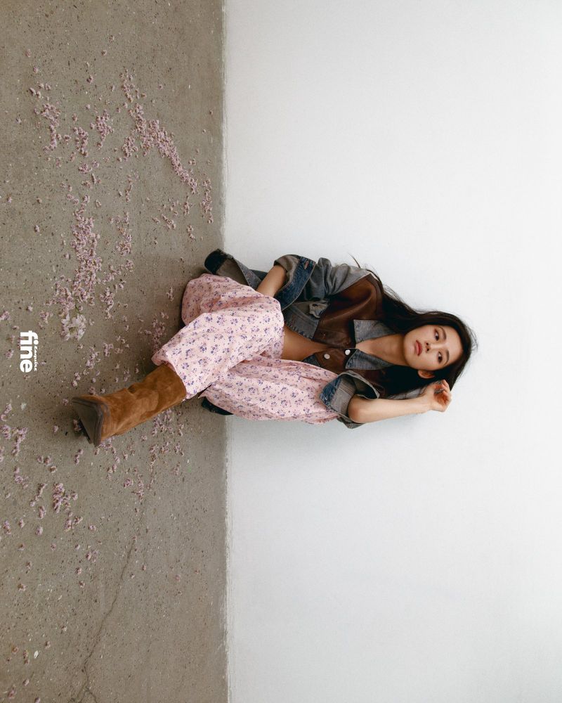 Young woman poses indoors with floral pants, jacket, and boots, lying on a grey and white floor next to the magazine 'fine'.