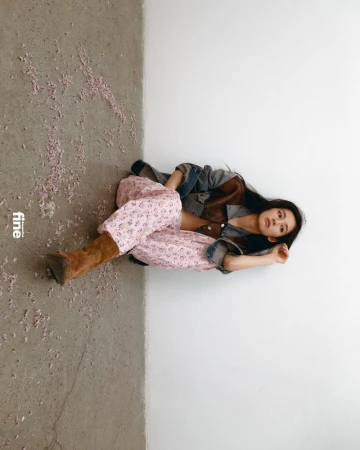 Young woman poses indoors with floral pants, jacket, and boots, lying on a grey and white floor next to the magazine 'fine'.