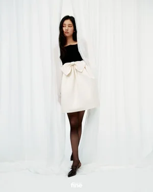 Woman in a black and cream bow mini dress and sheer tights standing amidst white sheer curtains.