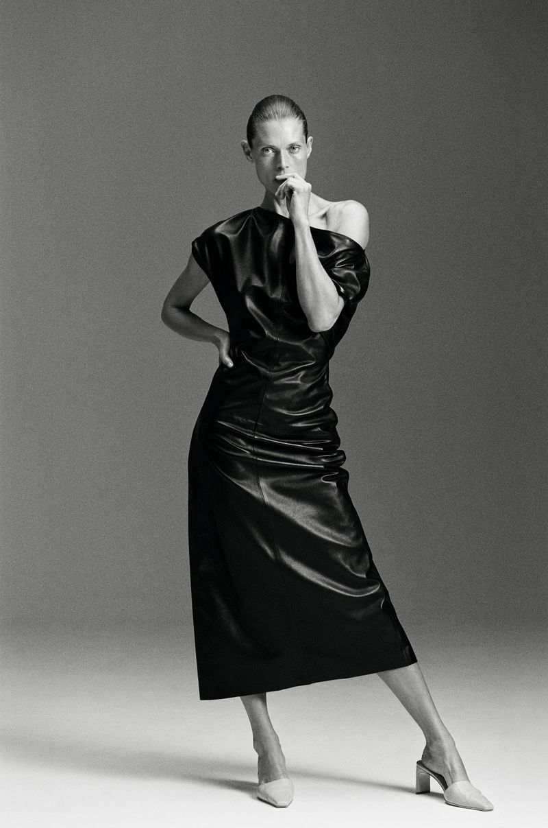 Malgosia Bela posing in a black off-the-shoulder midi dress and light pointed mules against a seamless grey backdrop.