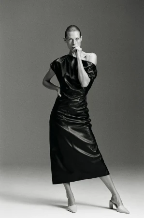 Malgosia Bela posing in a black off-the-shoulder midi dress and light pointed mules against a seamless grey backdrop.
