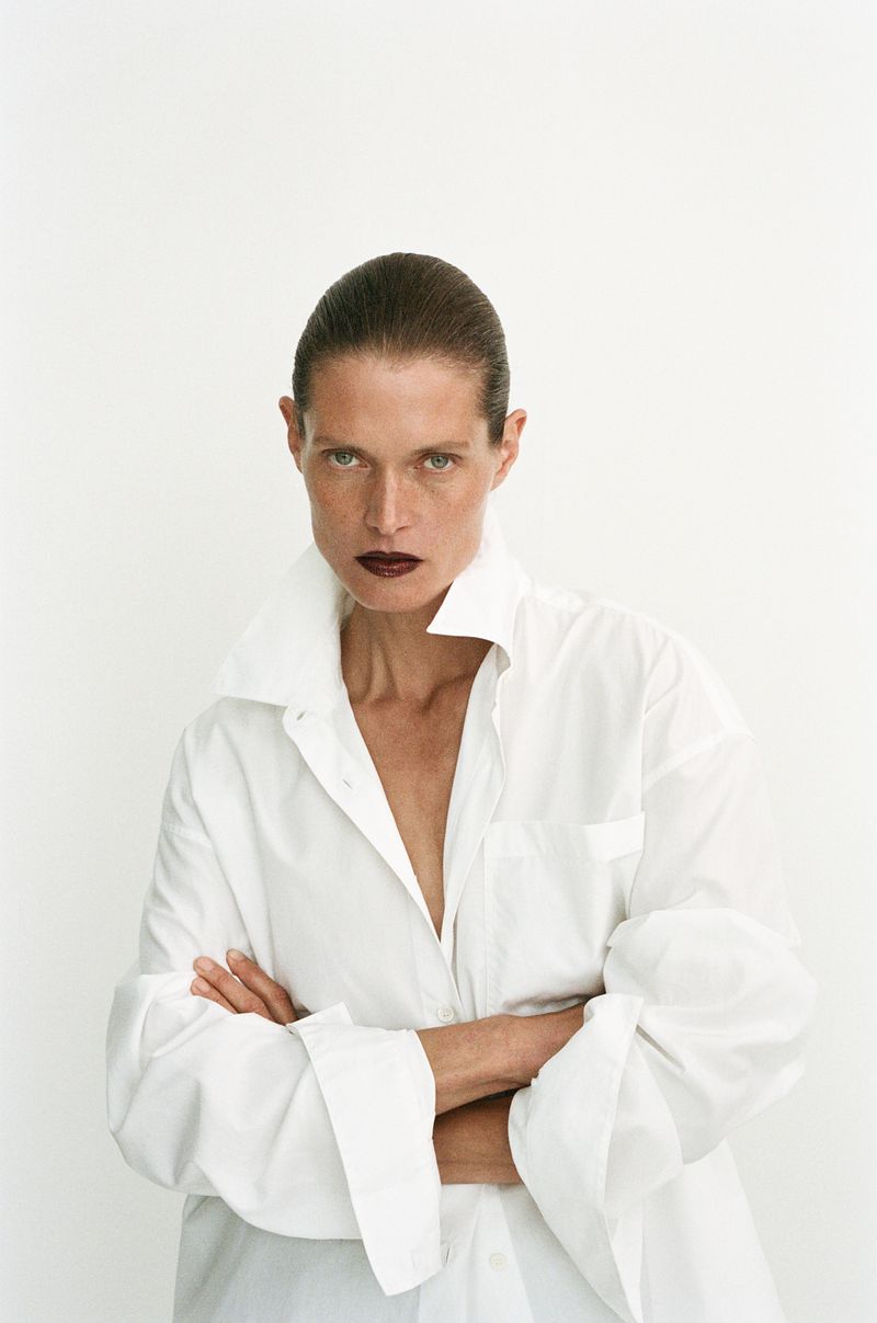 Model Malgosia Bela with dark hair and dark lipstick wearing an oversized white button-down shirt against a plain white background.