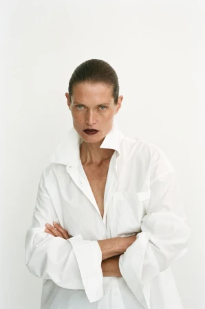 Model Malgosia Bela with dark hair and dark lipstick wearing an oversized white button-down shirt against a plain white background.