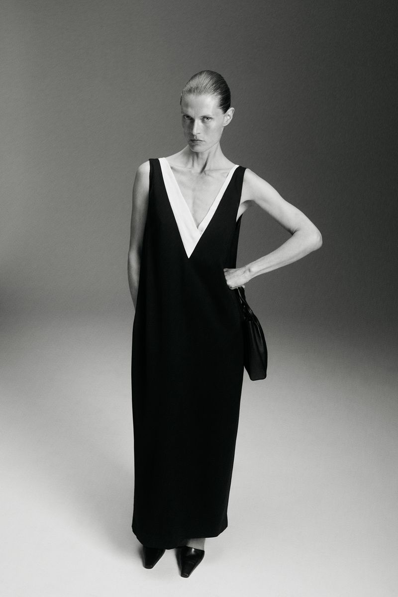 Model Malgosia Bela poses in a black sleeveless maxi dress with a deep white V-neck insert against a studio background.