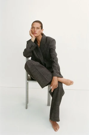 Malgosia Bela barefoot in a tailored charcoal grey suit seated on a metal chair against a white studio background.
