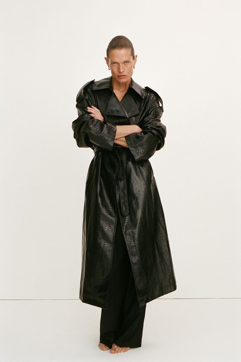 Model Malgosia Bela wears a long black leather trench coat with crossed arms against an off-white seamless background.