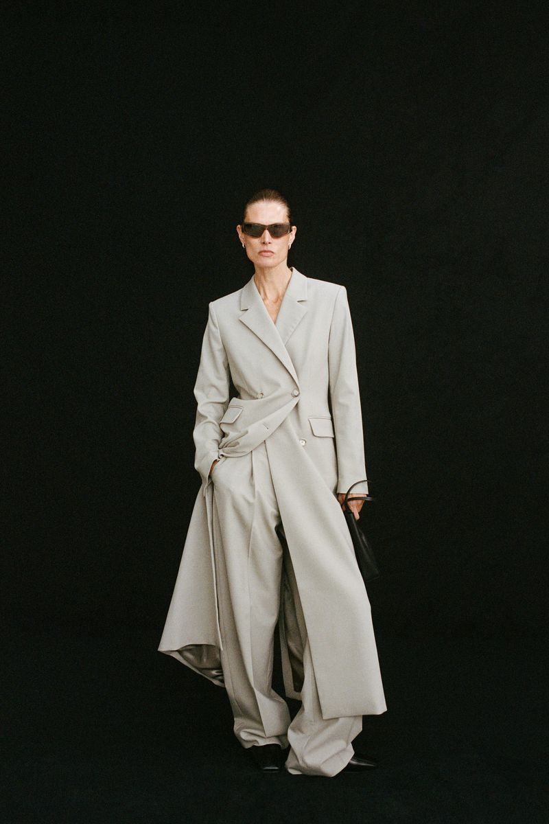 Malgosia Bela wearing an oversized light beige tailored trench suit with sunglasses against a solid black background.
