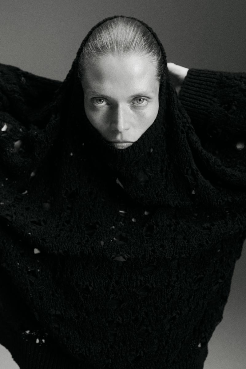 Monochrome portrait of Malgosia Bela wearing a high textured black knit sweater pulled over her nose against a studio backdrop.