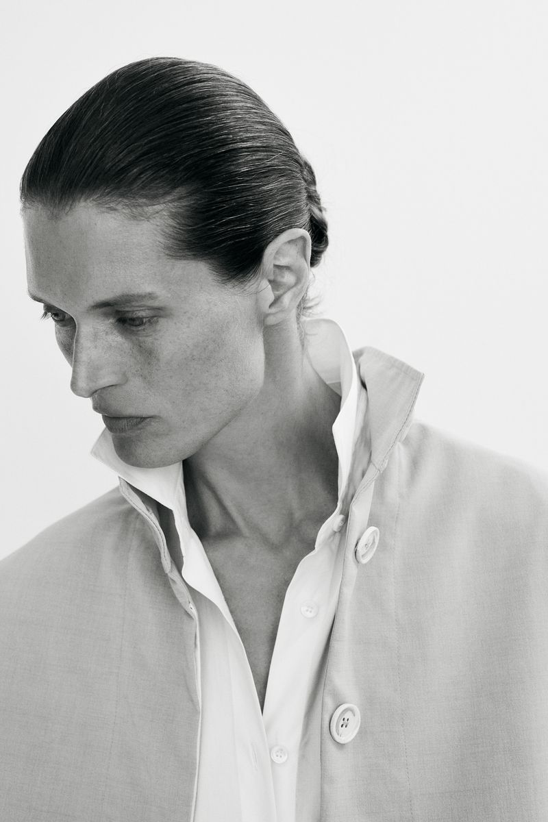 Black and white portrait of model Malgosia Bela looking down, wearing a high-collar coat and a white shirt against a plain background.