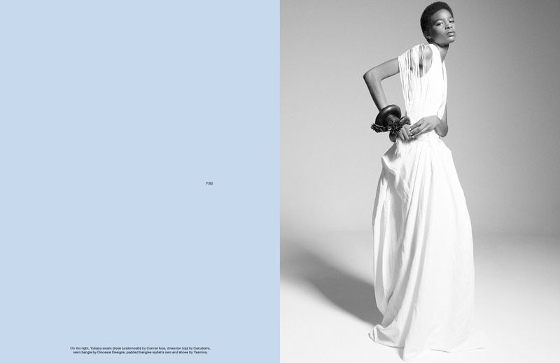 Black female model in white dress with large bangle in a studio with half blue background. Editorial fashion shot in black and white.
