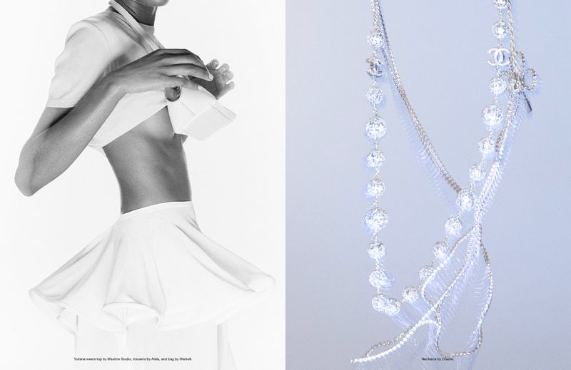 Black and white editorial fashion shot of a woman wearing a white skirt and top, accessorized with a bag and crystal necklace.