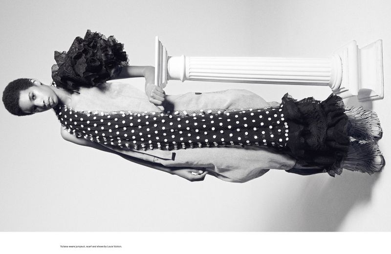 Black and white fashion shot of a person in a jumpsuit, polka dot scarf, and shoes by Louis Vuitton, posing with a white column.