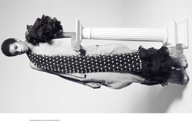 Model posing horizontally against a white studio background next to a classical pillar prop wearing polka dot clothing