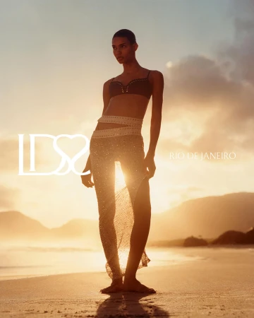 Model Barbara Valente stands barefoot on a sandy beach at sunset, wearing a dark bikini top and a sheer glittering sarong skirt.