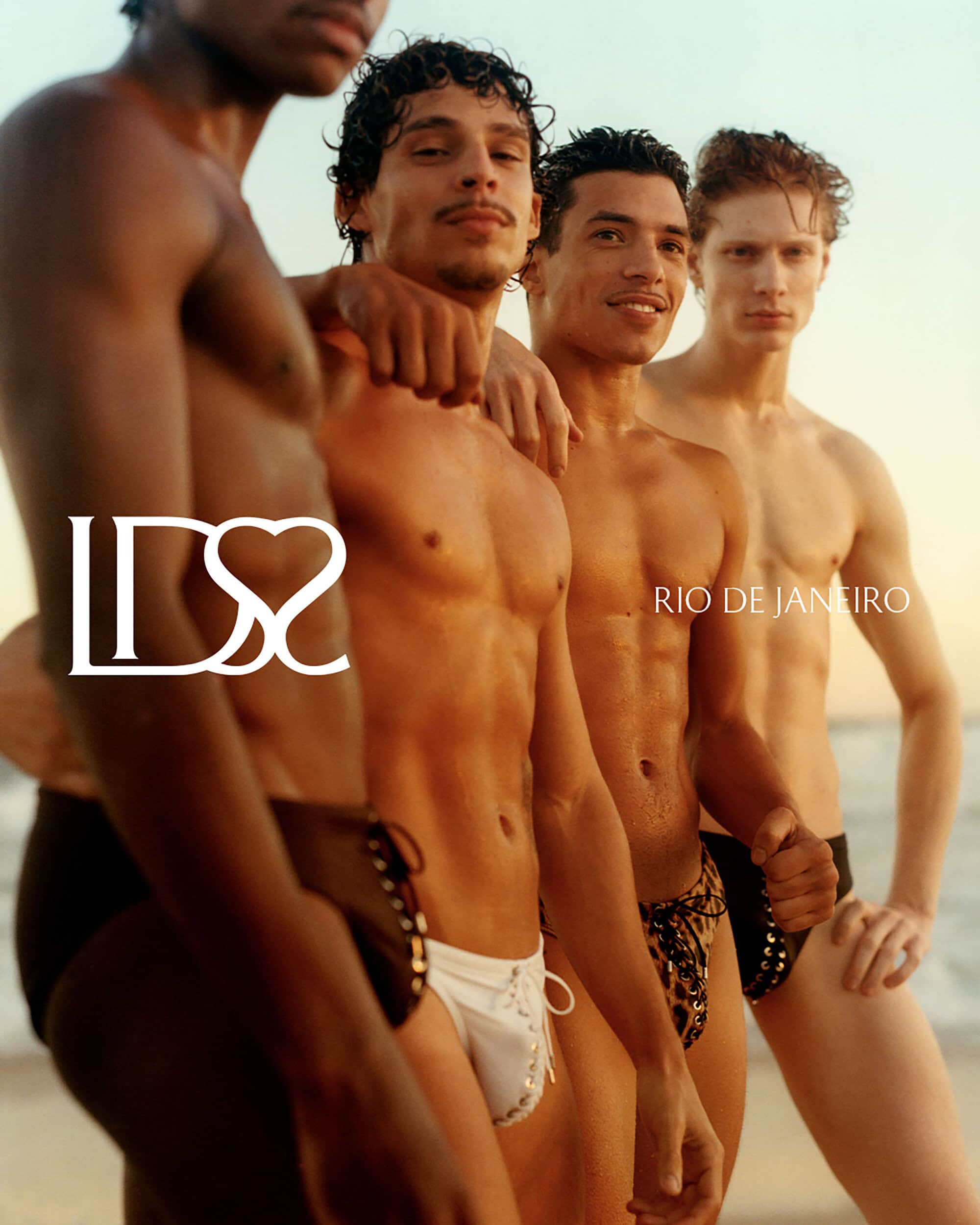 Four shirtless male models wearing swimwear posing together on a sandy beach at sunset for Ludovic de Saint Sernin.