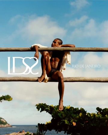 Model perched on a rustic wooden railing wearing black swimwear briefs against a blue sky for Ludovic de Saint Sernin in Rio de Janeiro.
