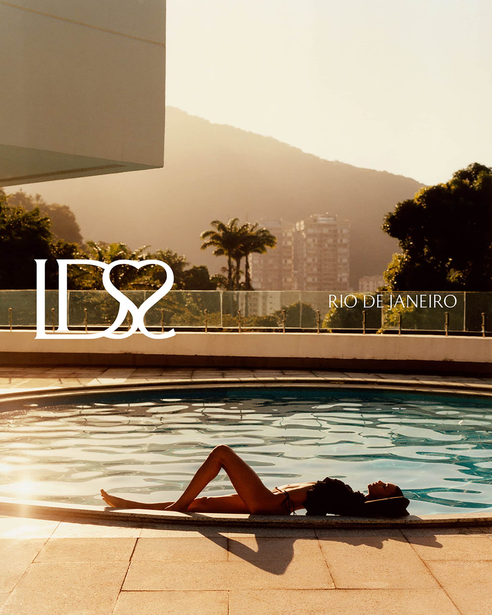 A model reclining beside an outdoor swimming pool during golden hour with palm trees and hills in the background.