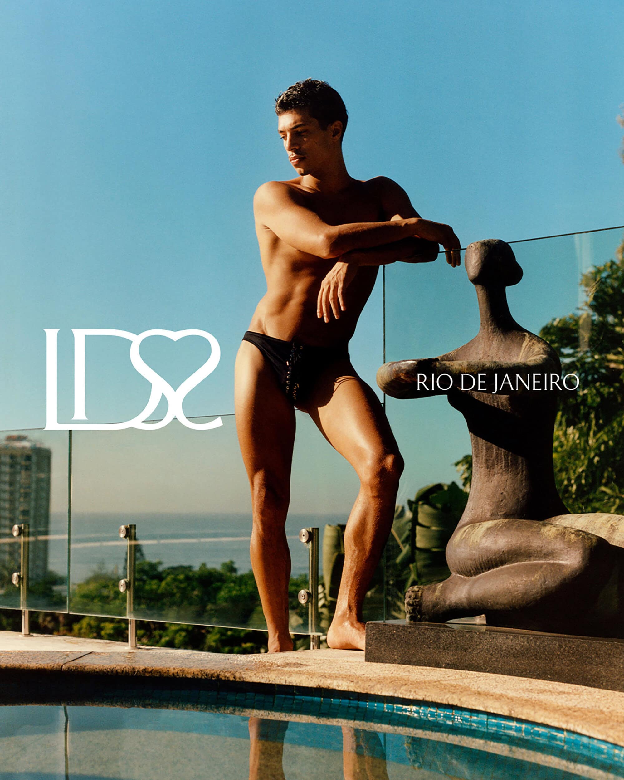 A man in black lace-up swim briefs poses beside a poolside sculpture overlooking the coast in Rio de Janeiro.