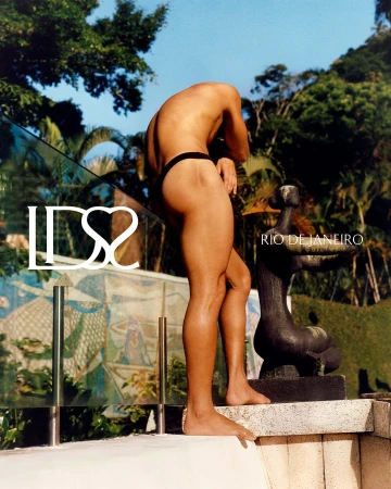 Man wearing black swimwear standing near a balcony with a dark sculptural figure against lush green foliage.