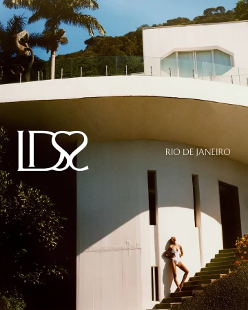 Model standing on outdoor steps against a white modernist building facade with palm trees and a bright sunny sky.