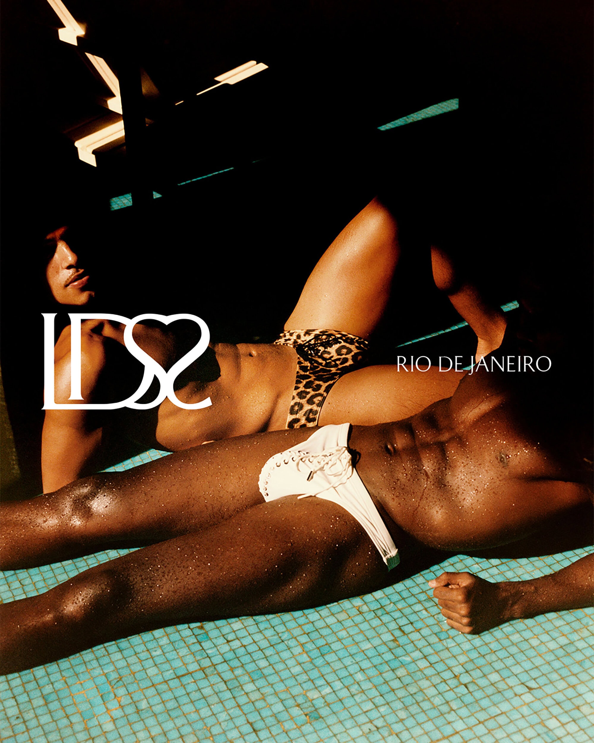 Two shirtless male models reclining in a tiled swimming pool wearing white lace-up and leopard print swim briefs under dramatic lighting.