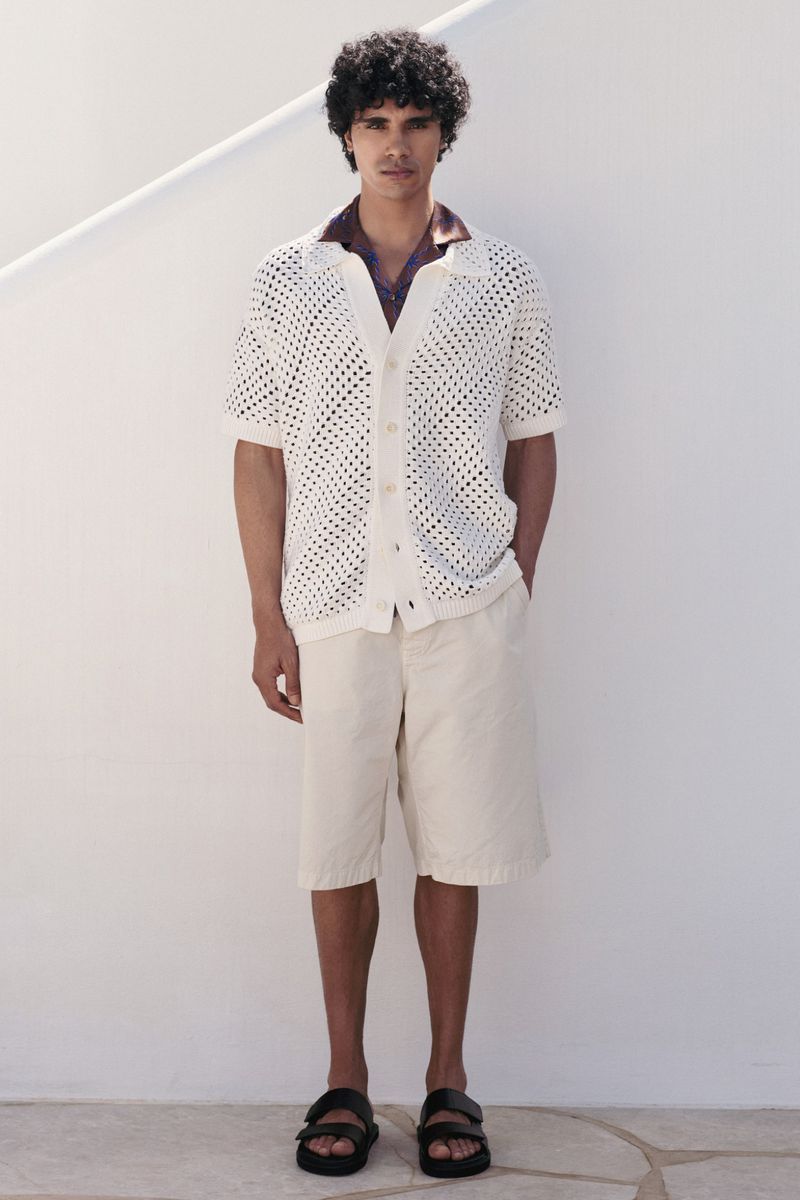 Model Kamal Sad in an off-white open-knit shirt, cream shorts, and black sandals standing against a white wall.
