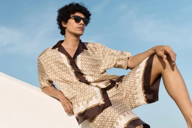 Model Kamal Sad in sunglasses and a patterned cream and brown silk shirt and matching shorts against a clear blue sky.