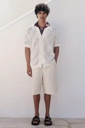 Model Kamal Sad in an off-white open-knit shirt, cream shorts, and black sandals standing against a white wall.