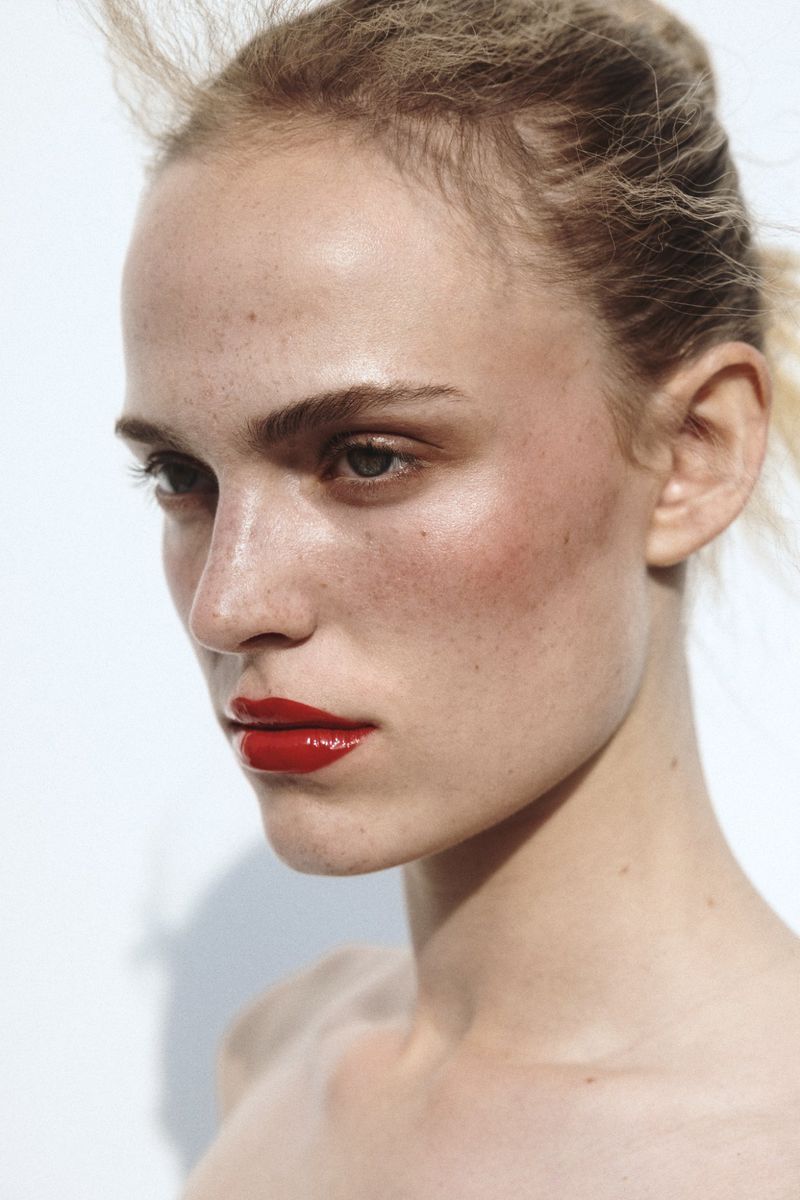 Close-up portrait of an adult woman with fair skin, freckles, and striking red lipstick, set against a clean white backdrop.