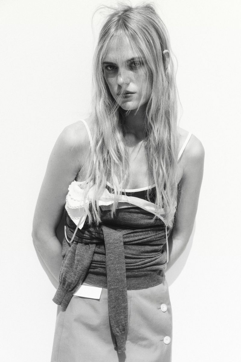 Monochrome fashion portrait of a slender adult woman with long blond hair wearing a layered sleeveless top and skirt against a white background.