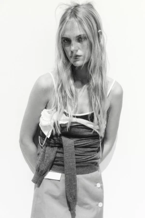 Monochrome fashion portrait of a slender adult woman with long blond hair wearing a layered sleeveless top and skirt against a white background.