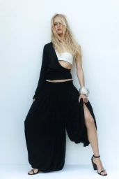 Model Rikke Nora Palsgaard Ronne in an asymmetric black top and voluminous wide-leg pants against a white studio background.