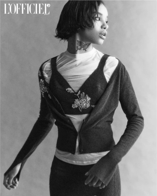 Black and white portrait of a young woman with a butterfly tattoo on her neck, wearing a dark cardigan and skirt, set against a plain backdrop.