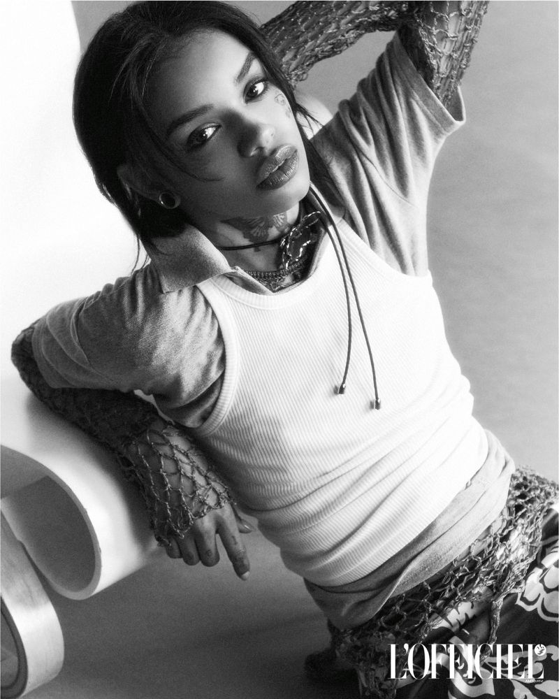 Monochrome portrait of an adult woman with patterned tattoos, wearing casual clothing, posing indoors against a light background with a chair.