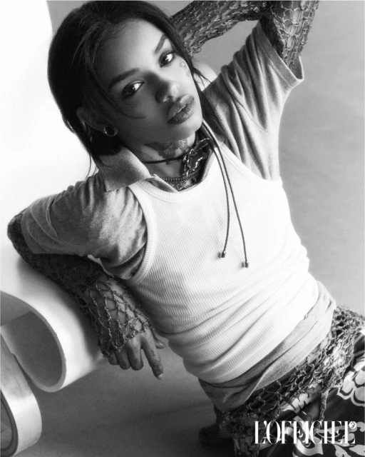 Monochrome portrait of an adult woman with patterned tattoos, wearing casual clothing, posing indoors against a light background with a chair.