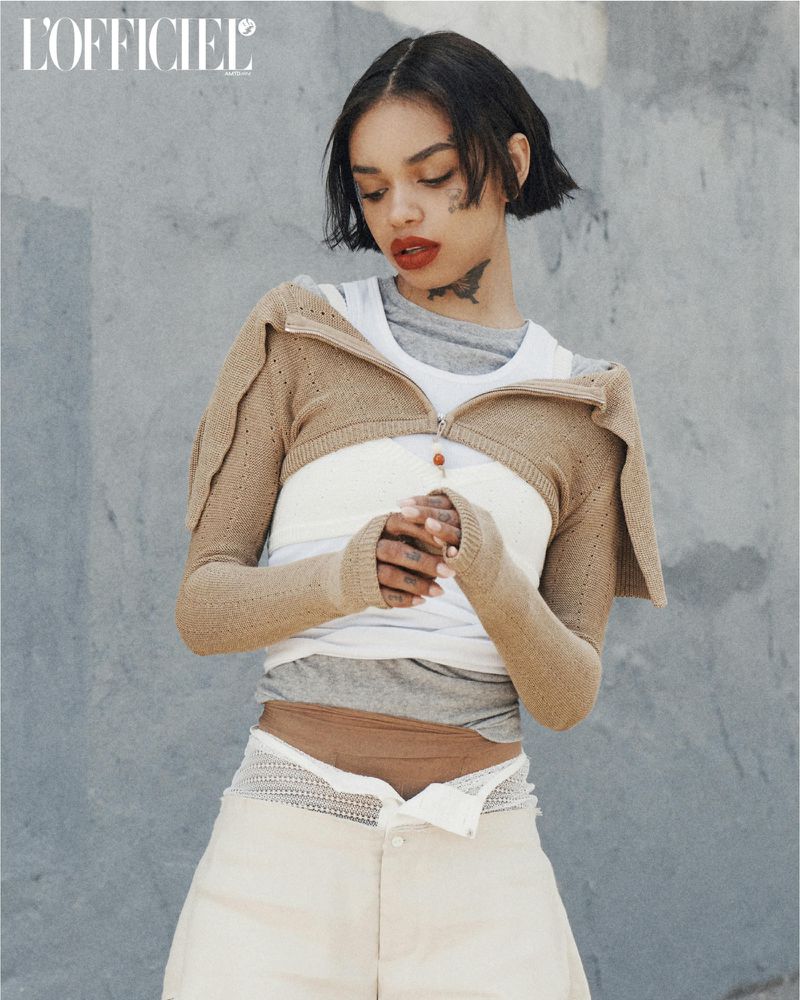Portrait of a woman with short dark hair wearing a knit sweater, white top, and light-colored shorts posing against a textured gray wall.