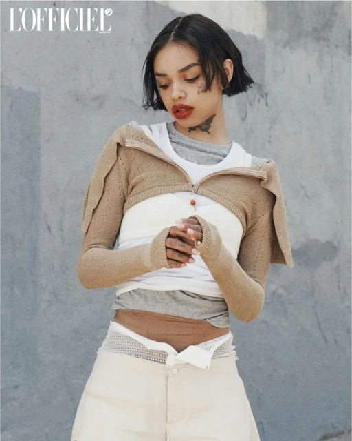 Portrait of a woman with short dark hair wearing a knit sweater, white top, and light-colored shorts posing against a textured gray wall.