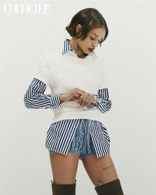 Fashion editorial shot of a tattooed model posing in a white sweater vest, striped shirt, and light-blue knitted shorts with brown suede boots.