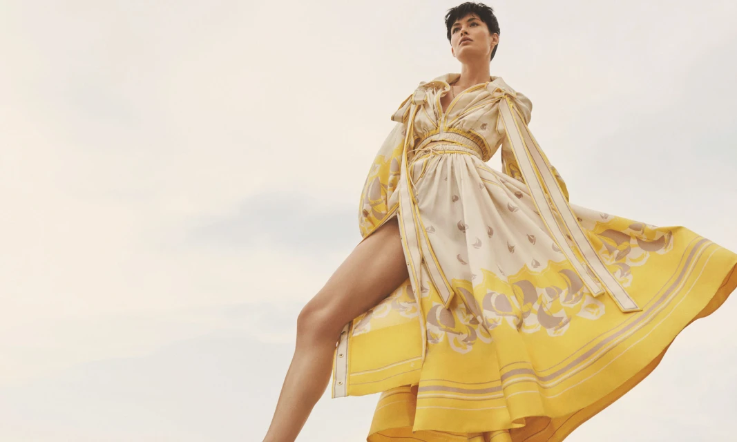 Elegant woman in a flowing yellow and white patterned dress posing against a soft, overcast sky, showcasing high fashion apparel.