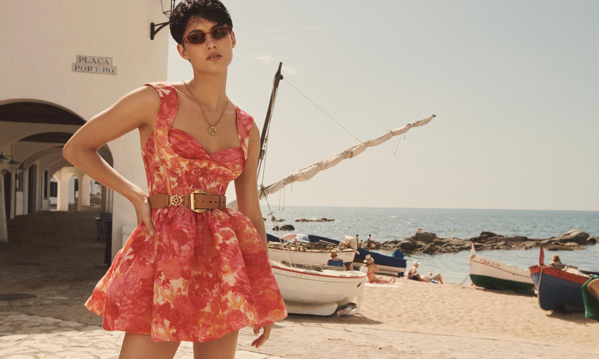 Woman in a floral sundress with belt, posing on a sandy beach near fishing boats with the ocean and a white building in the background.