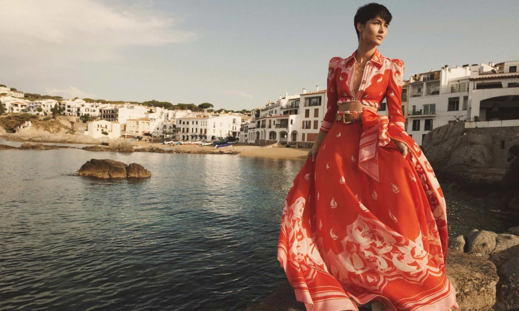 An adult female model poses in an orange patterned dress on a rocky outcrop, overlooking a white coastal town and the sea.