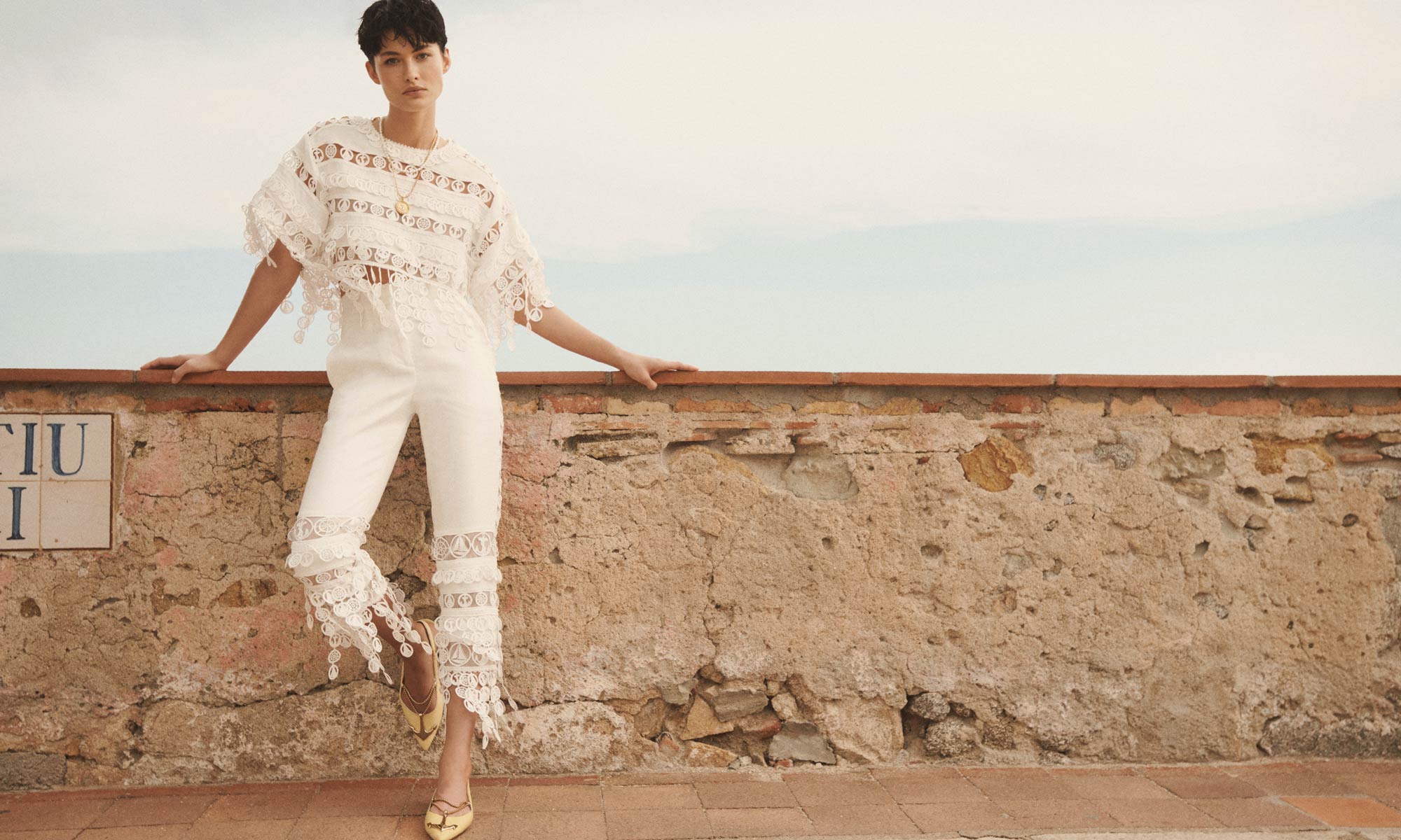 A fashionable woman in a white lace top and cropped pants poses confidently against a weathered wall under a bright, overcast sky.