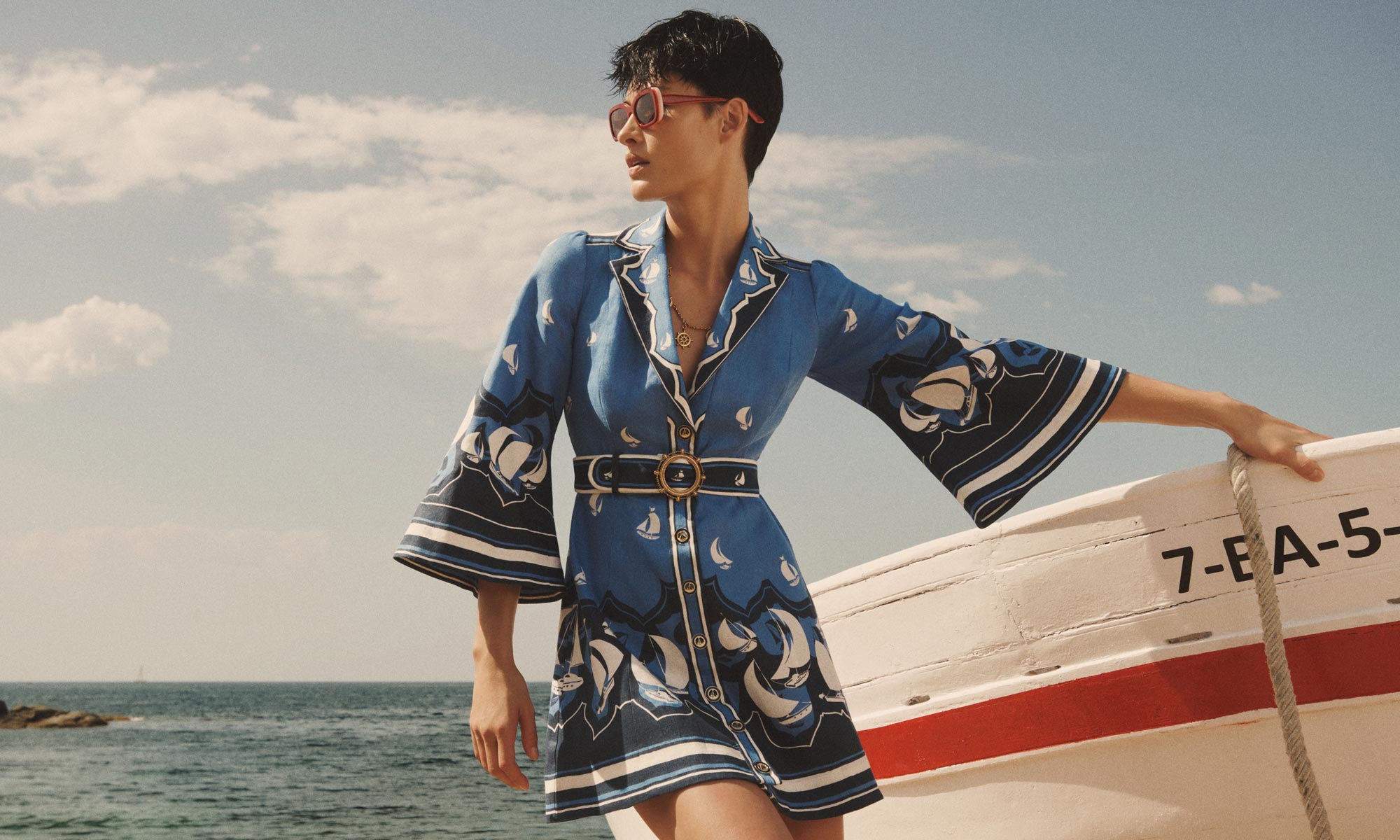 Model wearing a blue patterned dress and sunglasses leaning against a white boat by the ocean under a clear sky.